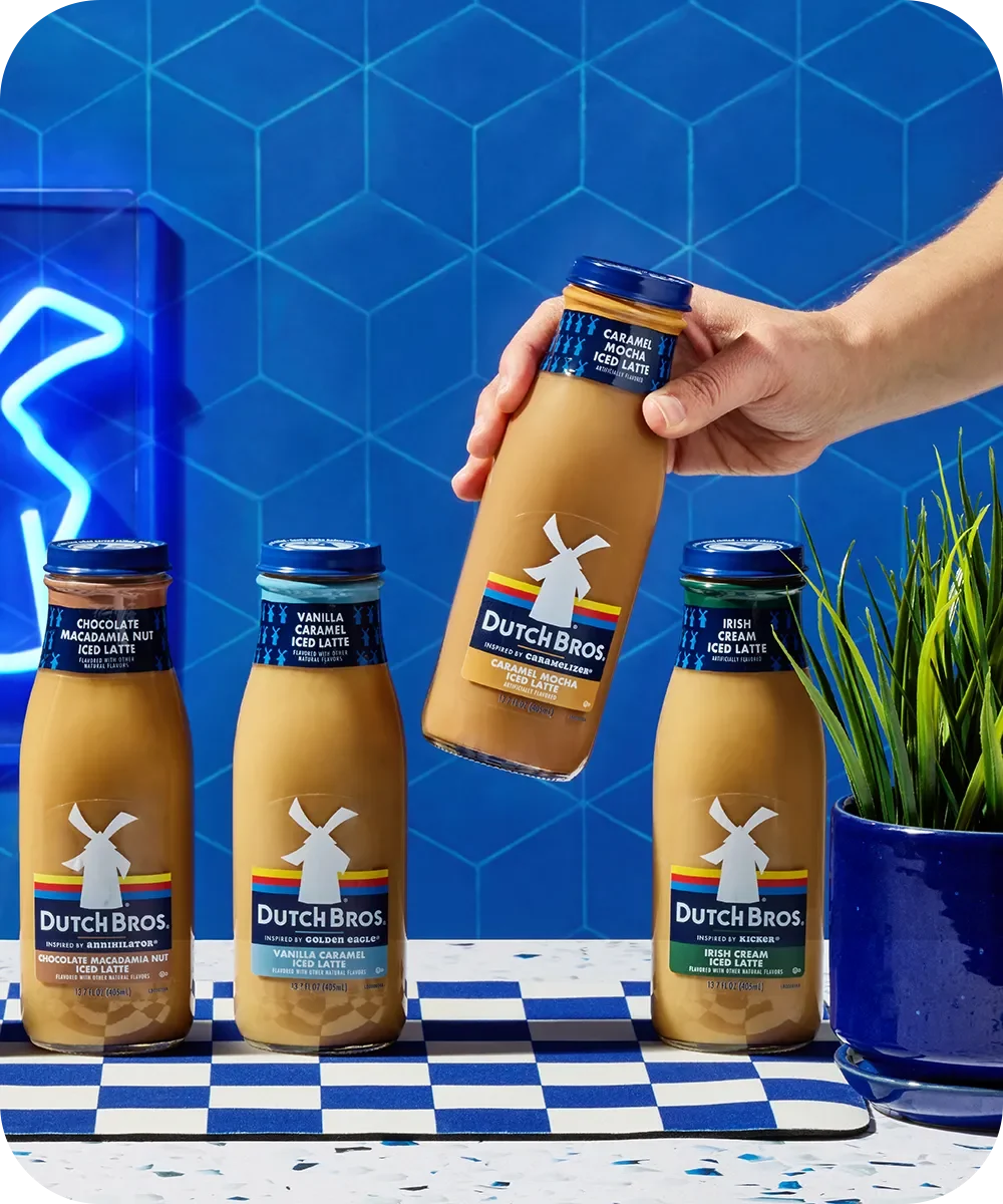Hand holding Dutch Bros Caramel Mocha Iced Latte bottle above three other flavored coffee drinks against blue tiled background.