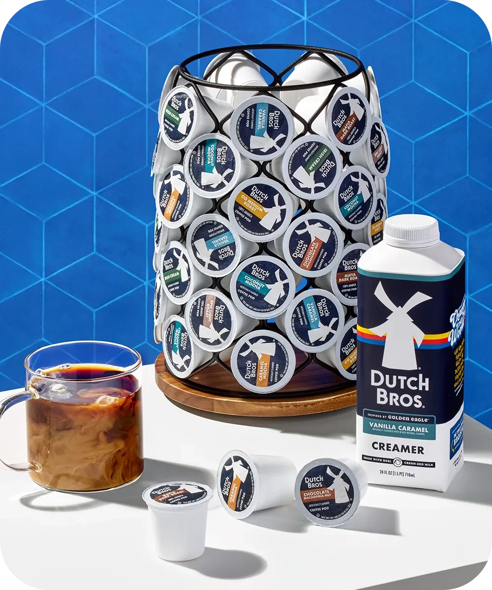 Dutch Bros coffee pods in storage rack with creamer bottle and glass of iced coffee on blue geometric background.