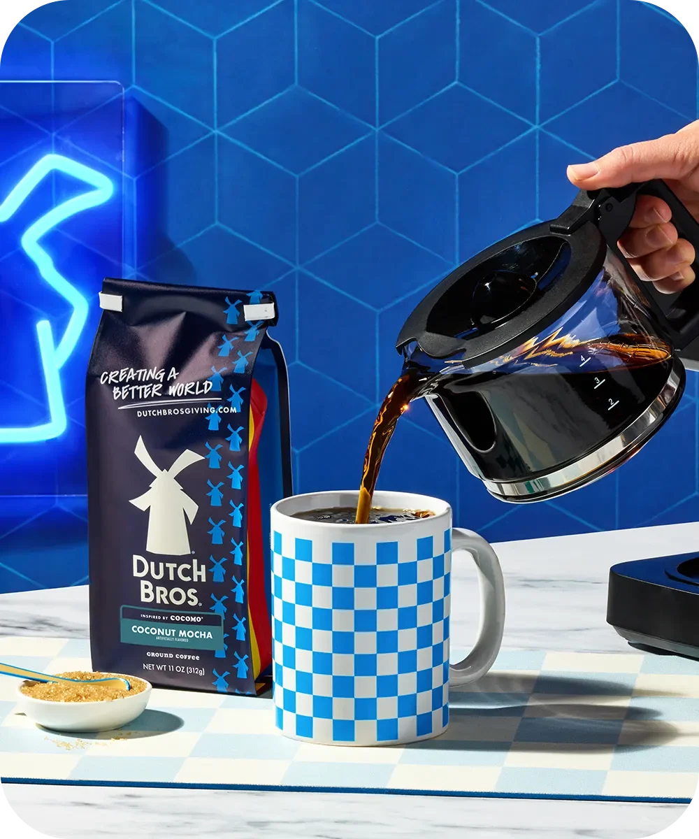 Hand pouring coffee from carafe into blue checkered mug, with Dutch Bros Coconut Mocha coffee bag against blue background.