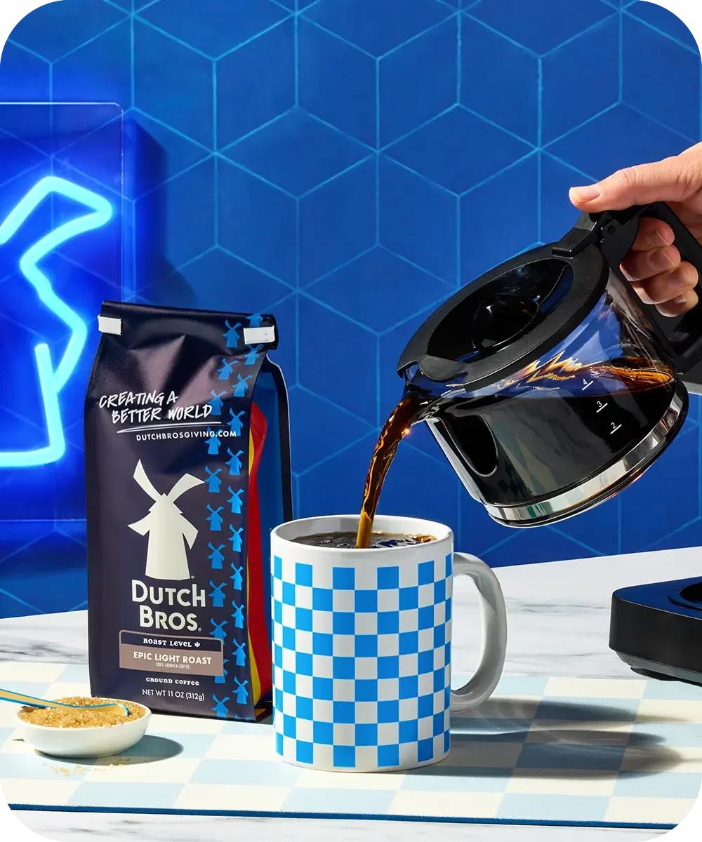 Hand pouring coffee from carafe into blue checkered mug next to Dutch Bros coffee bag against geometric blue background.