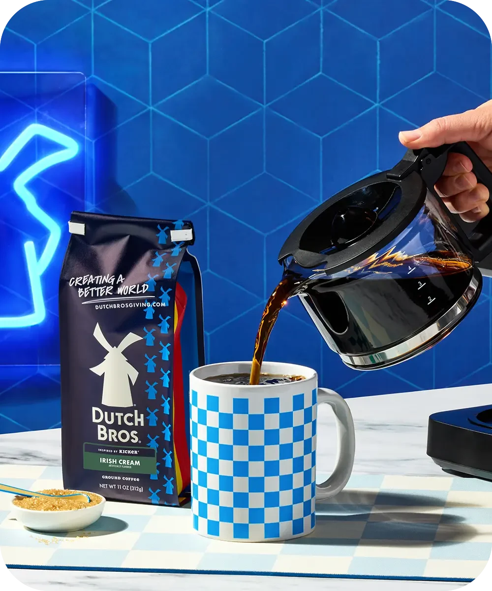 Hand pouring coffee from carafe into blue checkered mug, with Dutch Bros coffee bag against blue geometric background.