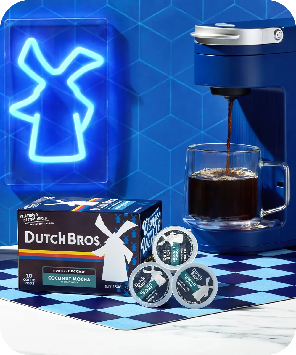 Blue coffee machine brewing into glass cup with Dutch Bros Coconut Mocha pods and box against blue windmill backdrop.