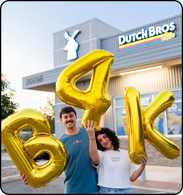 Two people holding large gold letter balloons spelling "B4K" outside a Dutch Bros Coffee shop with its windmill logo visible.