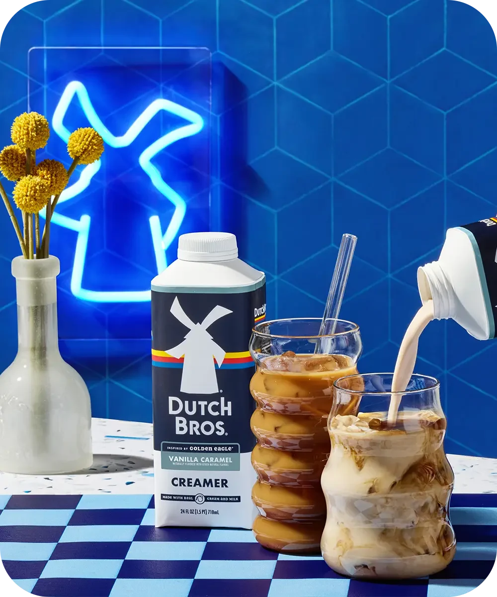 Dutch Bros creamer being poured into iced coffee drinks with blue neon sign and yellow flowers in background.
