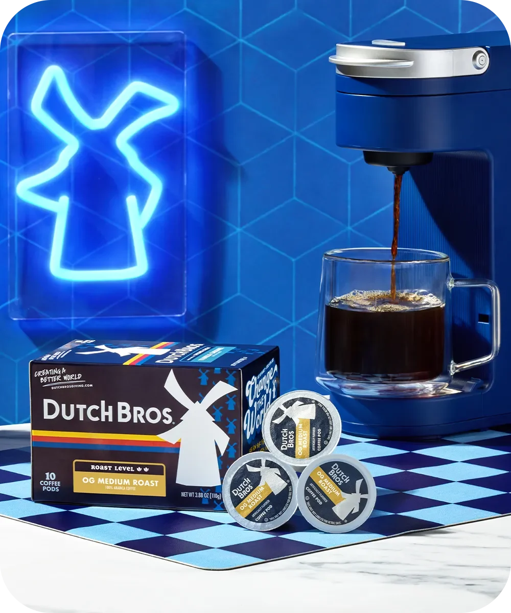 Dutch Bros coffee pods and machine brewing coffee into glass mug with blue neon windmill sign in background.