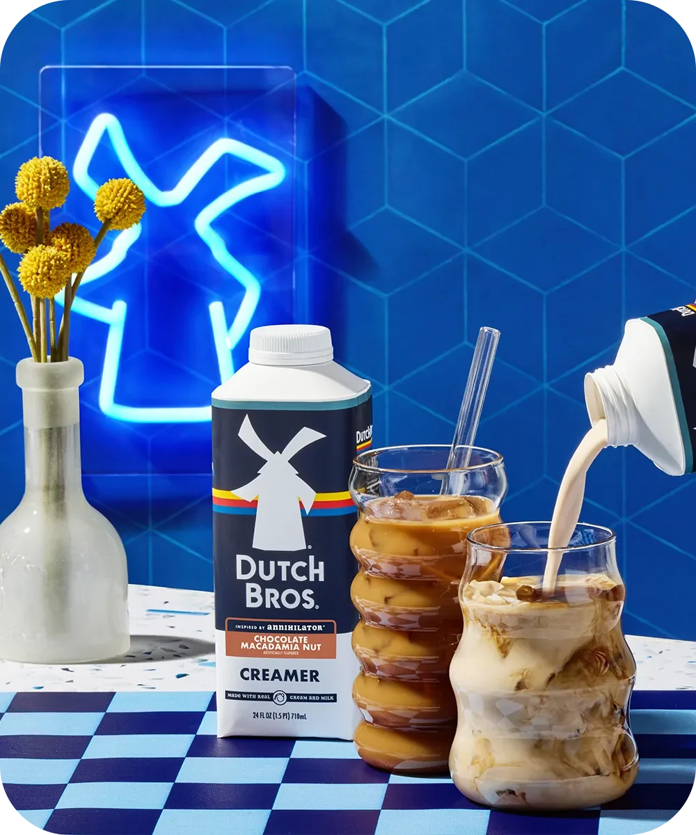 Dutch Bros creamer beside iced coffee drinks on checkered surface with blue neon sign and yellow flowers.