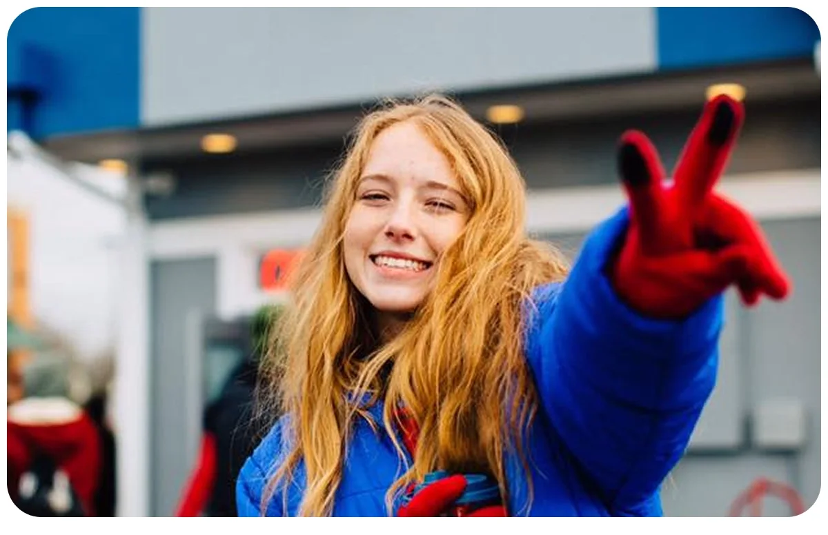 Person with long blonde hair wearing a bright blue coat and red gloves making a peace sign gesture outdoors.
