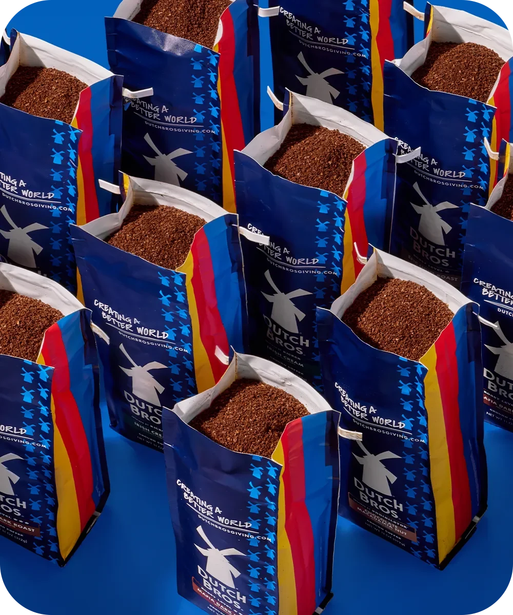 Several blue Dutch Bros coffee bags with windmill logos and colorful stripes containing ground coffee against blue background.