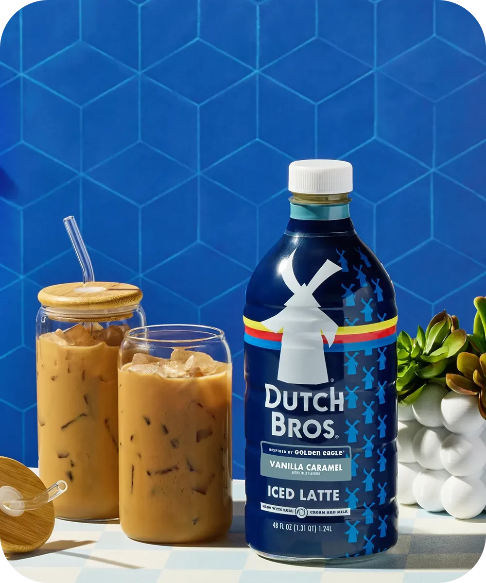 Dutch Bros Vanilla Caramel Iced Latte bottle with two glasses of iced coffee against blue hexagonal tile background.