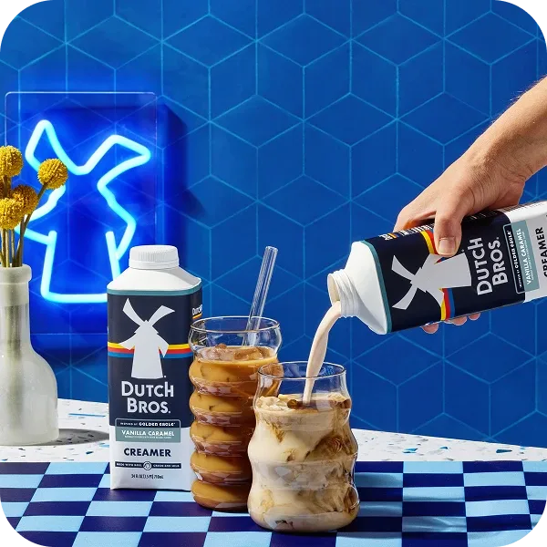 Hand pouring Dutch Bros. creamer into iced coffee drink with stacked donuts against blue geometric background with neon windmill sign.
