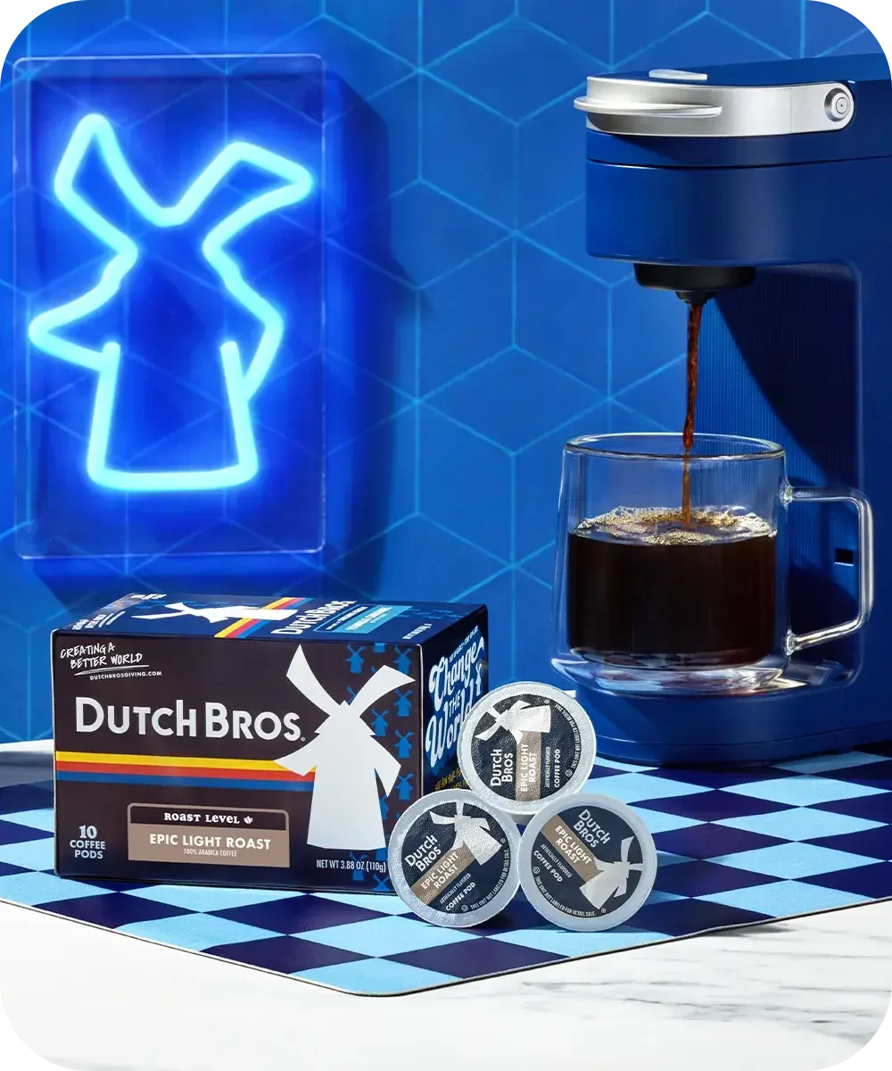 Blue coffee setup with Dutch Bros box, coffee pods, brewing machine pouring into glass cup, and neon windmill logo.