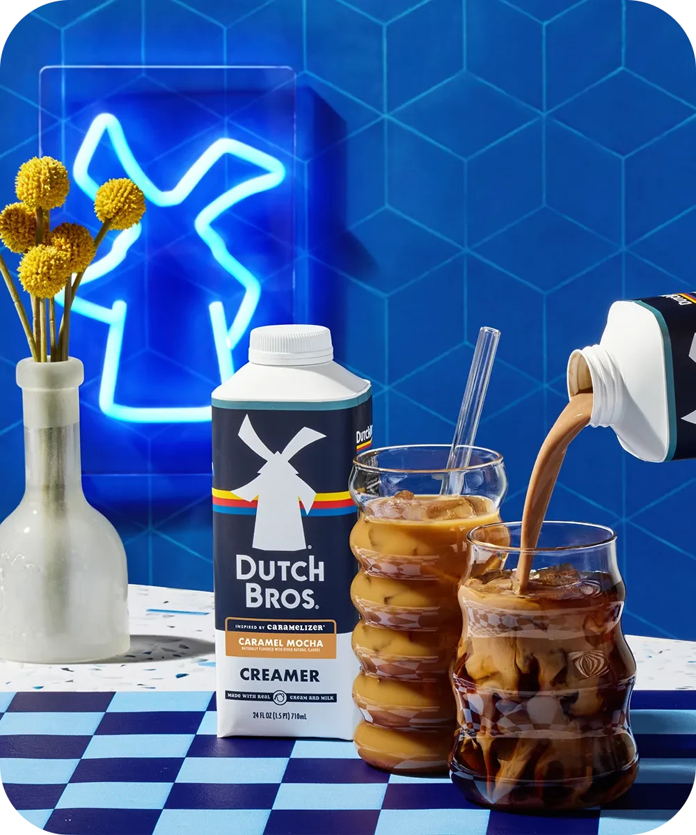 Dutch Bros Caramel Mocha creamer being poured into iced coffee with blue neon light and yellow flowers in background.