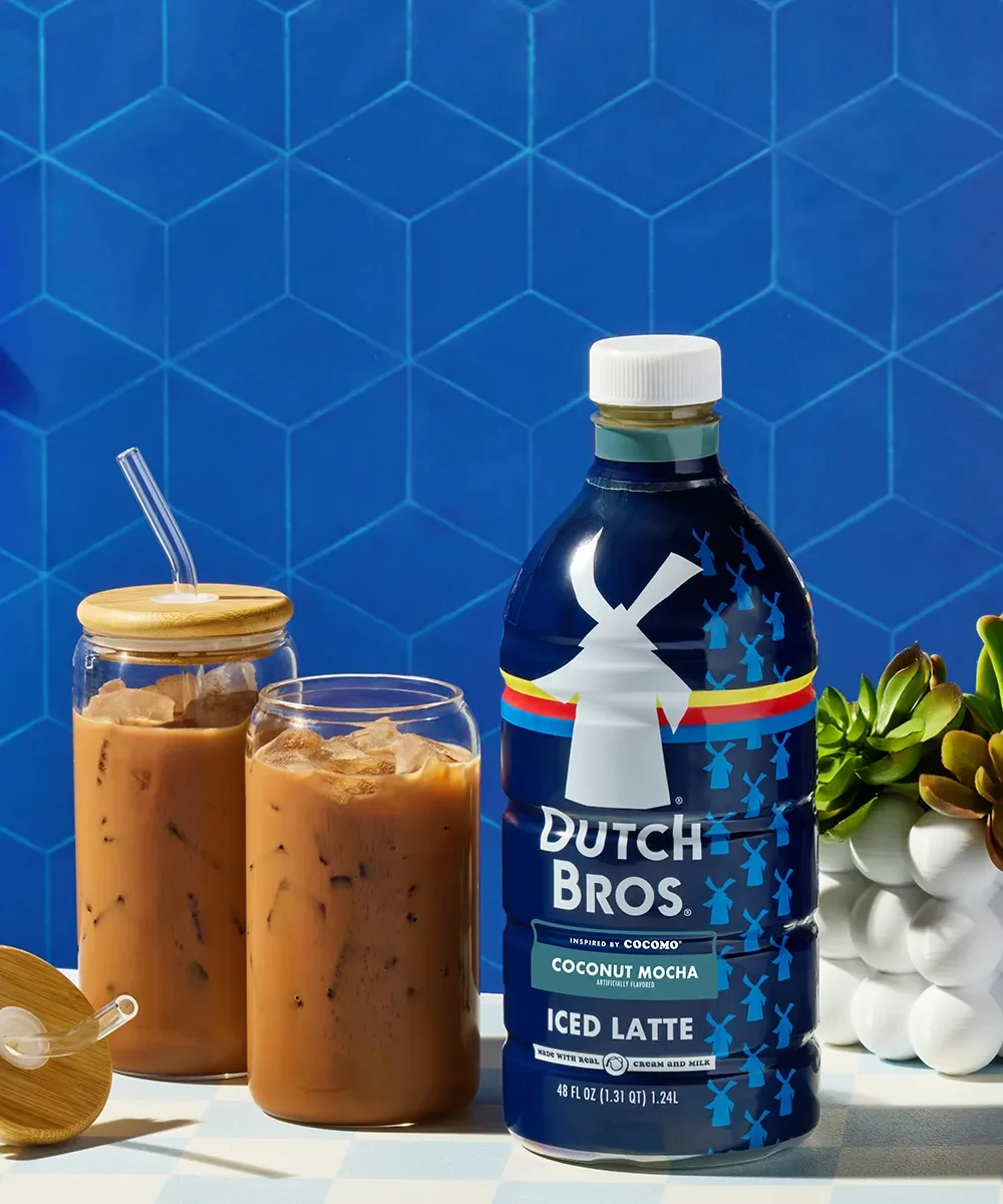 Dutch Bros Coconut Mocha Iced Latte bottle next to two glasses of iced coffee against blue hexagon tile background.