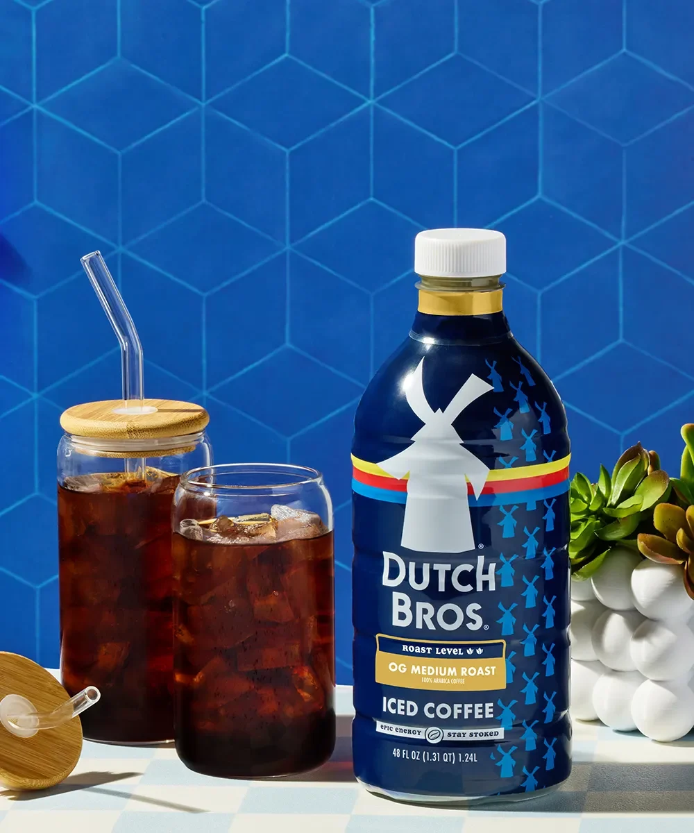 Dutch Bros iced coffee bottle next to glass jars of cold brew with ice against a blue hexagon-patterned background.